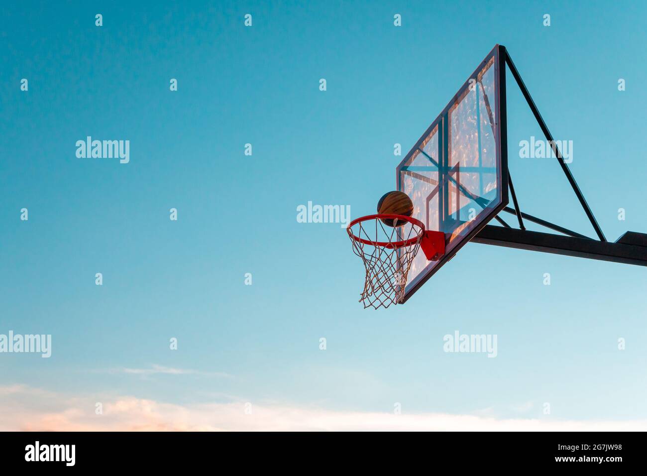 Ein Basketballkorb mit einem Ball auf einem blauen Himmel Hintergrund. Transparentes Kunststoff-Basketballschild auf dem Outdoor-Basketballfeld. Stockfoto