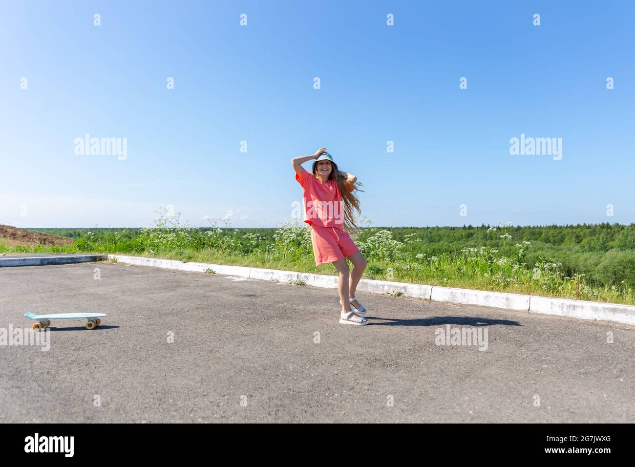 Ein lachendes Teenager-Mädchen mit einem Skateboard in einem trendigen Sommer-Anzug und einem panama-Hut mit flatterndem blondem Haar am blauen Himmel. Gesunder Lebensstil Stockfoto