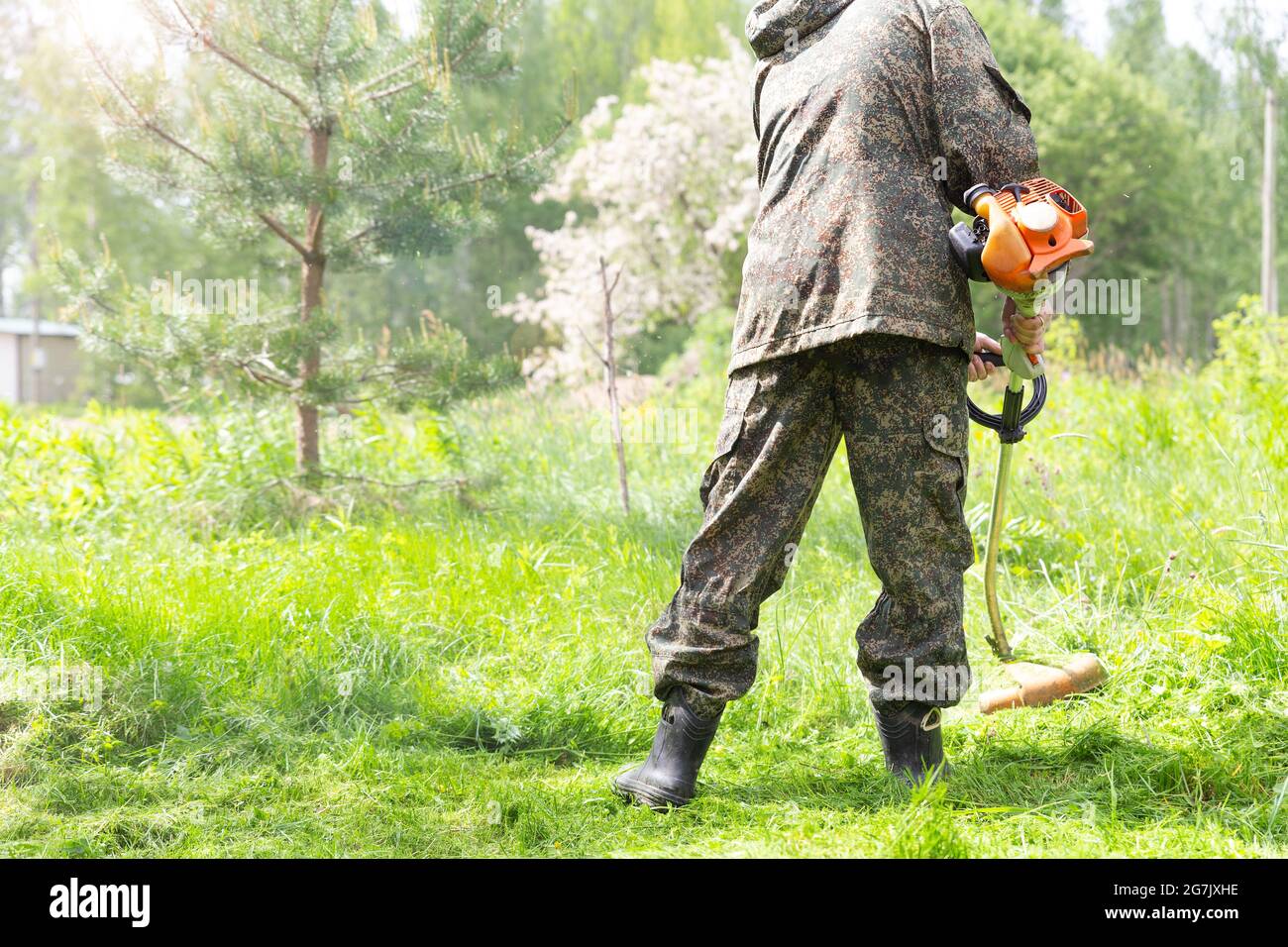 Ein Arbeiter in der Wohnmobil-Arbeitskleidung mäht hohes Gras mit Benzin-Rasentrimmer oder Rasenmäher im Stadtpark, Garten oder Hinterhof. Gartenpflege-Tools und Stockfoto
