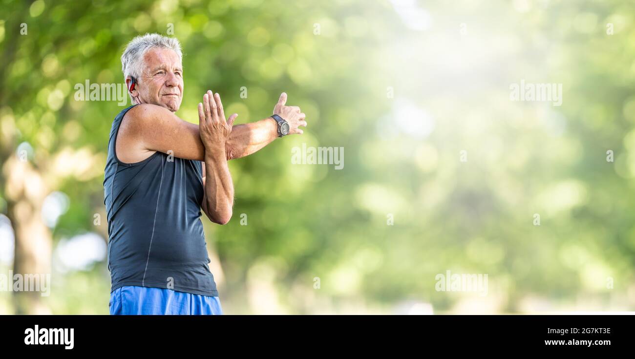 Ein älterer, flauer Mann streckt an einem Sommertag im Freien den Arm und trägt Uhren und Kopfhörer. Stockfoto