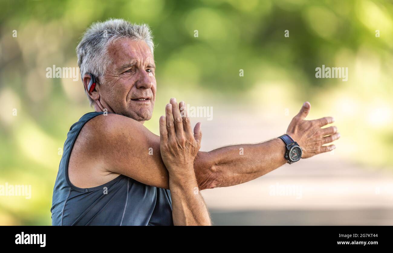 Ein älterer, flauer Mann streckt an einem Sommertag im Freien den Arm und trägt Uhren und Kopfhörer. Stockfoto