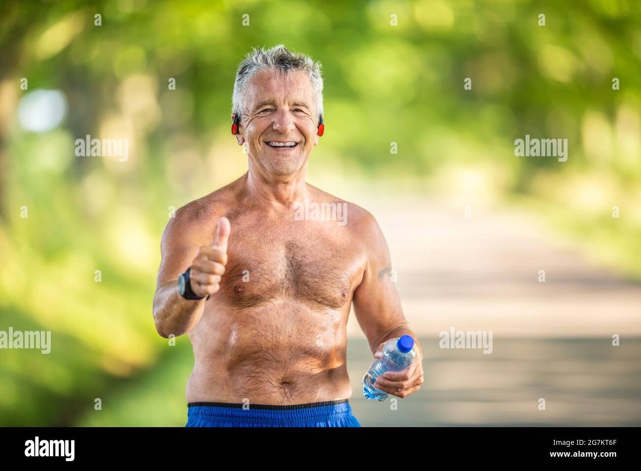 Der grauhaarige ältere Mann zeigt den Daumen nach oben, während er Wasser in Flaschen hält und nach einem Training in der Natur kein T-Shirt trägt. Stockfoto