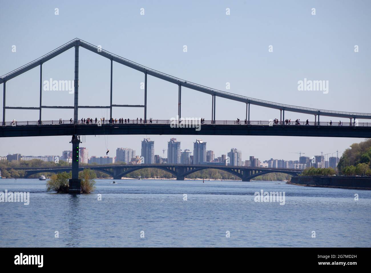 Parkovy Fußgängerbrücke über den Fluss Dnjepr mit Schiffen unter, Kiew, Ukraine. Silhouetten von Menschen, die auf der Brücke spazieren. Brücke bunjee ju Stockfoto