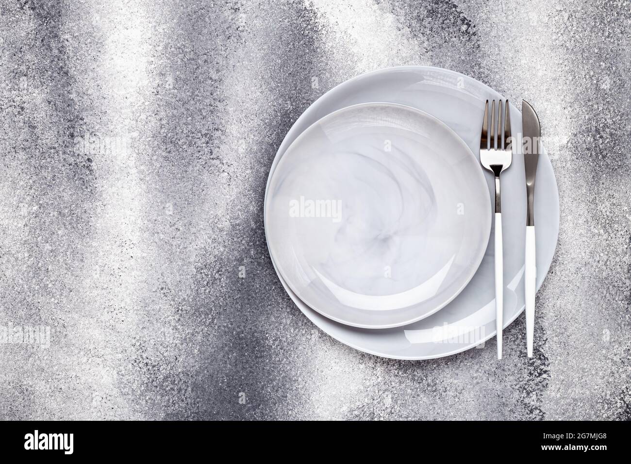 Zwei leere Keramikplatten und unbenutztes Messer mit Gabel, Geschirr auf einem grauen Grunge-Tisch, Servierkonzept. Flaches Lay-Design. Café-Menü. Grußkarte, Lay Stockfoto