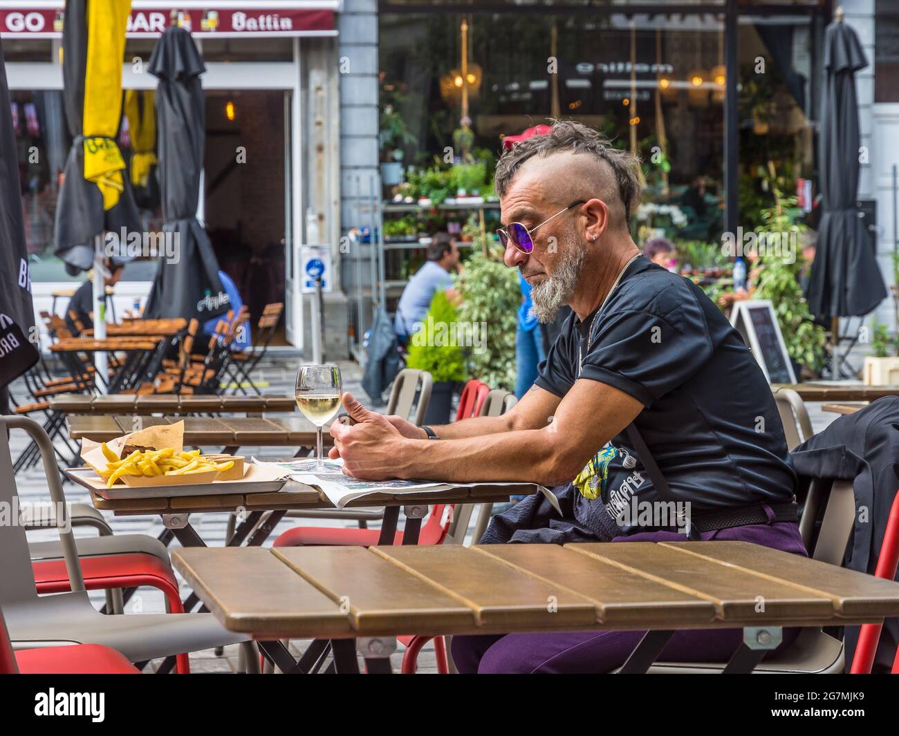 Älterer Mann im Punk-Stil, der Zeitung liest und Chips am Restauranttisch im Café im Freien in Saint Gilles, Brüssel, Belgien, isst. Stockfoto
