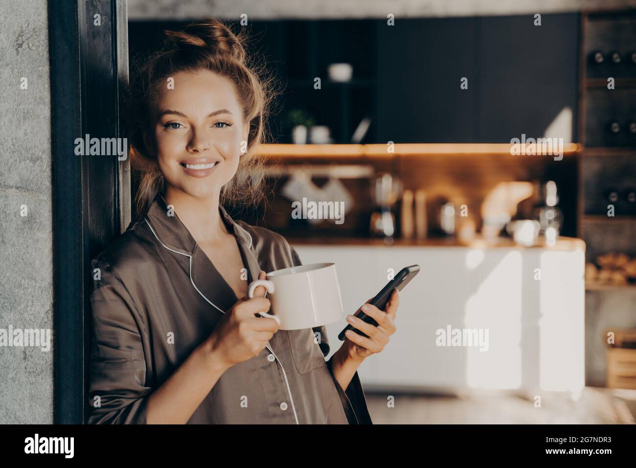 Schöne glückliche junge Frau, die ihren Morgenkaffee hat, während sie neue E-Mails und Benachrichtigungen auf dem Smartphone checkt Stockfoto