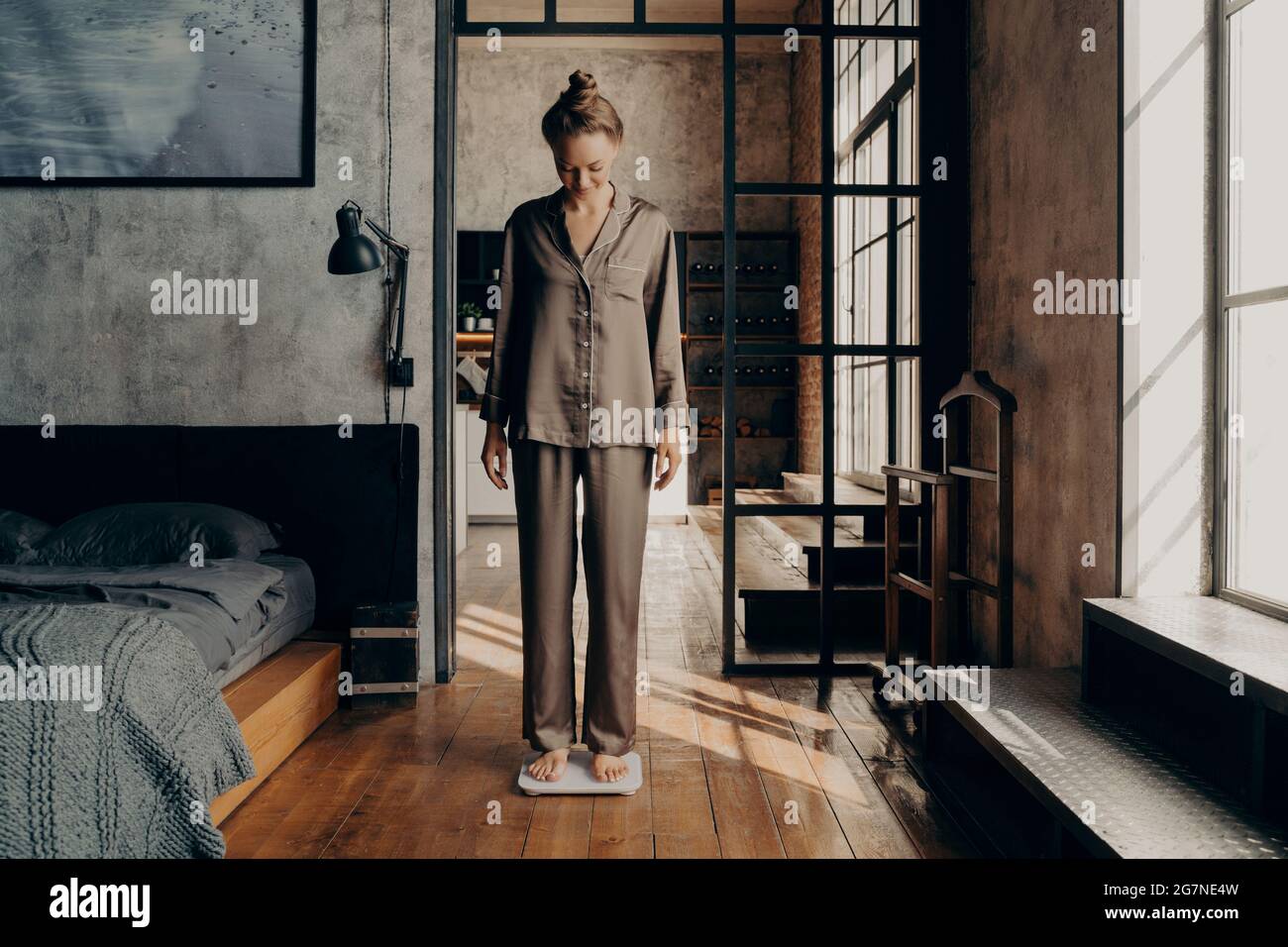Junge schöne Frau in bequemen Satin-Pyjama auf elektronische Waage im Schlafzimmer stehen Stockfoto