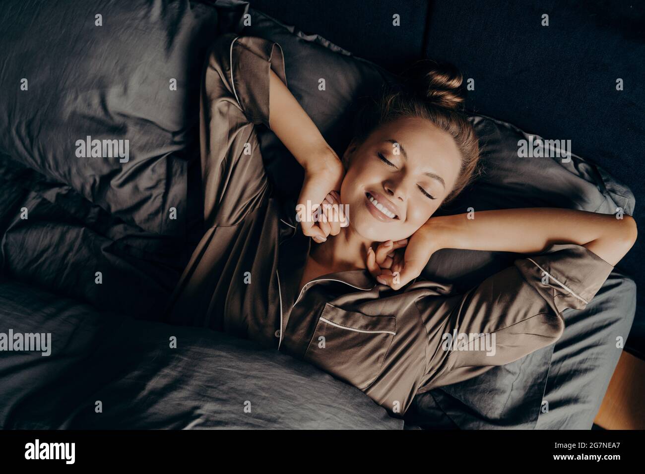 Junge schöne entspannte Brünette weiblich in Satin Pyjama Stretching im Bett Stockfoto