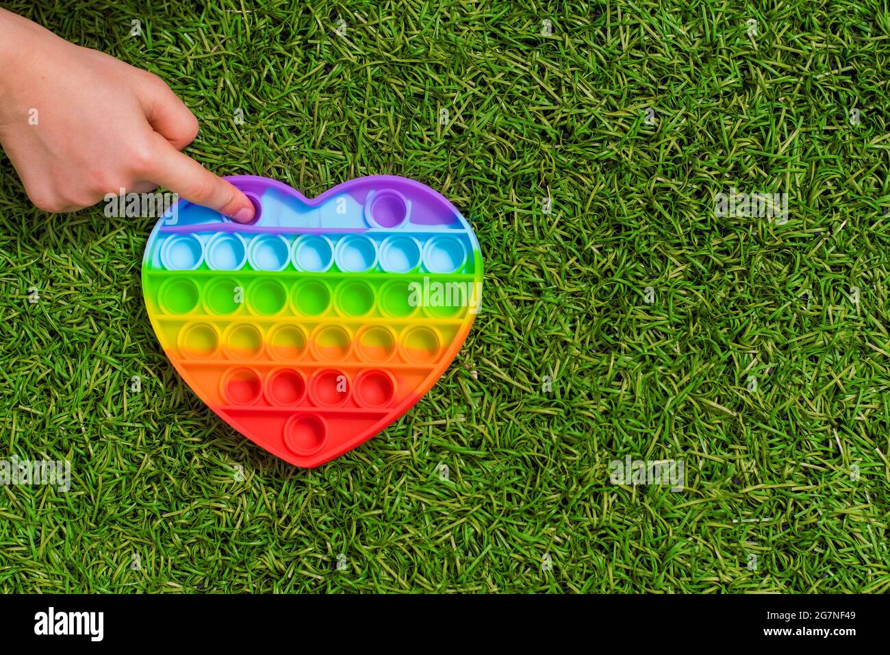 Ein Kind spielt mit einem bunten Regenbogen Anti-Stress Sinnesspielzeug Pop es in Form eines Herzens auf dem Gras. Kinderhand platzt Kugeln auf ein Spielzeug Pop it in Stockfoto