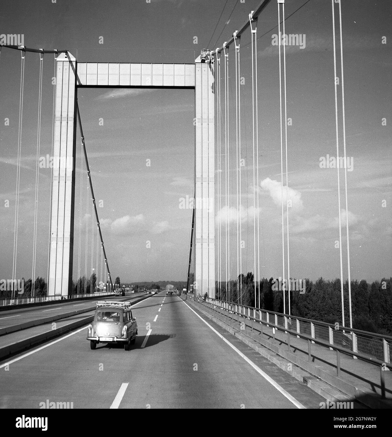 1960er, historisch, Deutschland, ein Auto der damaligen Zeit, das auf einer neu gebauten Hängebrücke unterwegs war. Seit Anfang 1800 werden solche Brücken mit Aufhängeseilen an vertikalen Hosenträgern eingesetzt, um das Deck, den tragenden Abschnitt, an dem der Verkehr sich kreuzt, zu stützen. Stockfoto