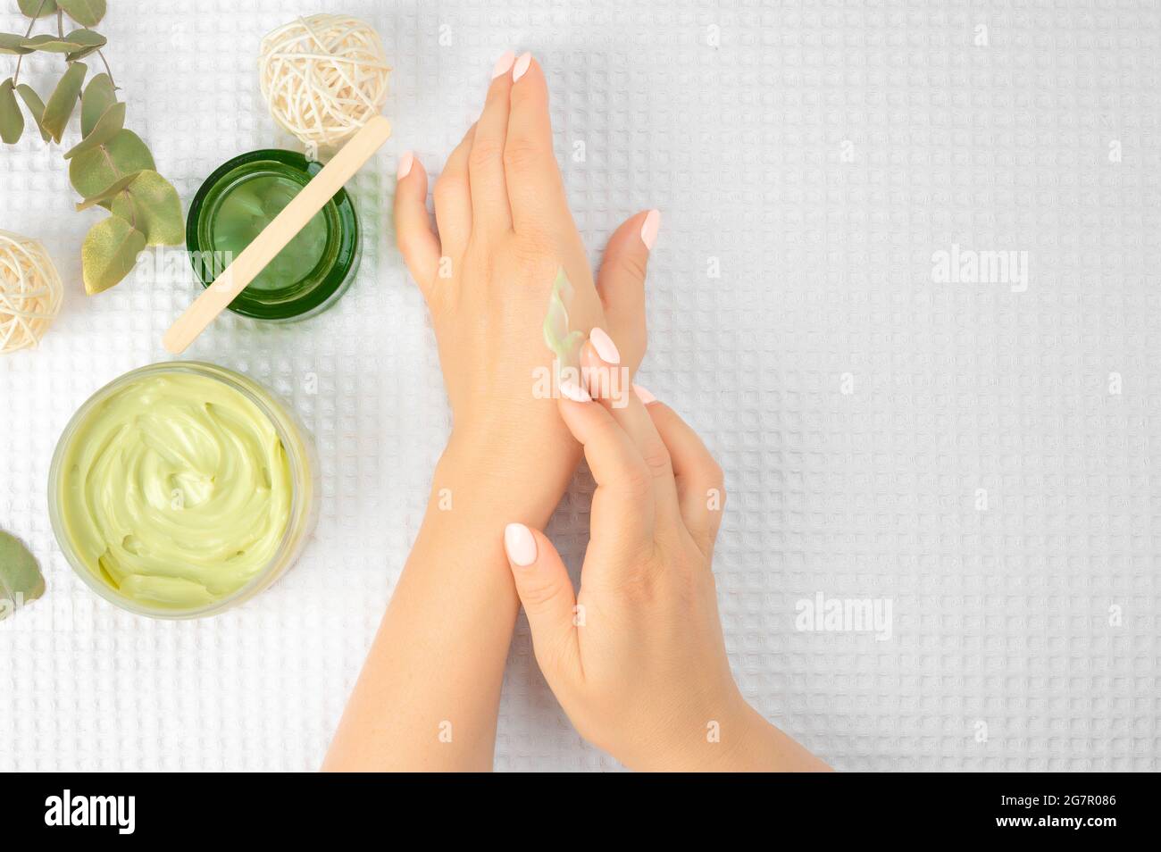 Frauenhand mit Aloe Vera-Gel, Avocadoöl, Creme auf dem Finger über dem Glas mit kosmetischer Feuchtigkeitscreme und grünen Blättern auf weißem Hintergrund, flaches Lay-Comp Stockfoto