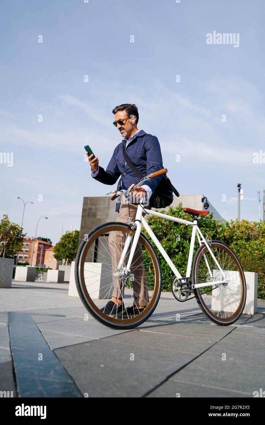 Erwachsener Mann, der mit einer Hand das Fahrrad hält und mit der anderen Hand auf sein Mobiltelefon schaut, zum Büro. Umweltfreundliches Transportkonzept. Hochwertige Fotos Stockfoto