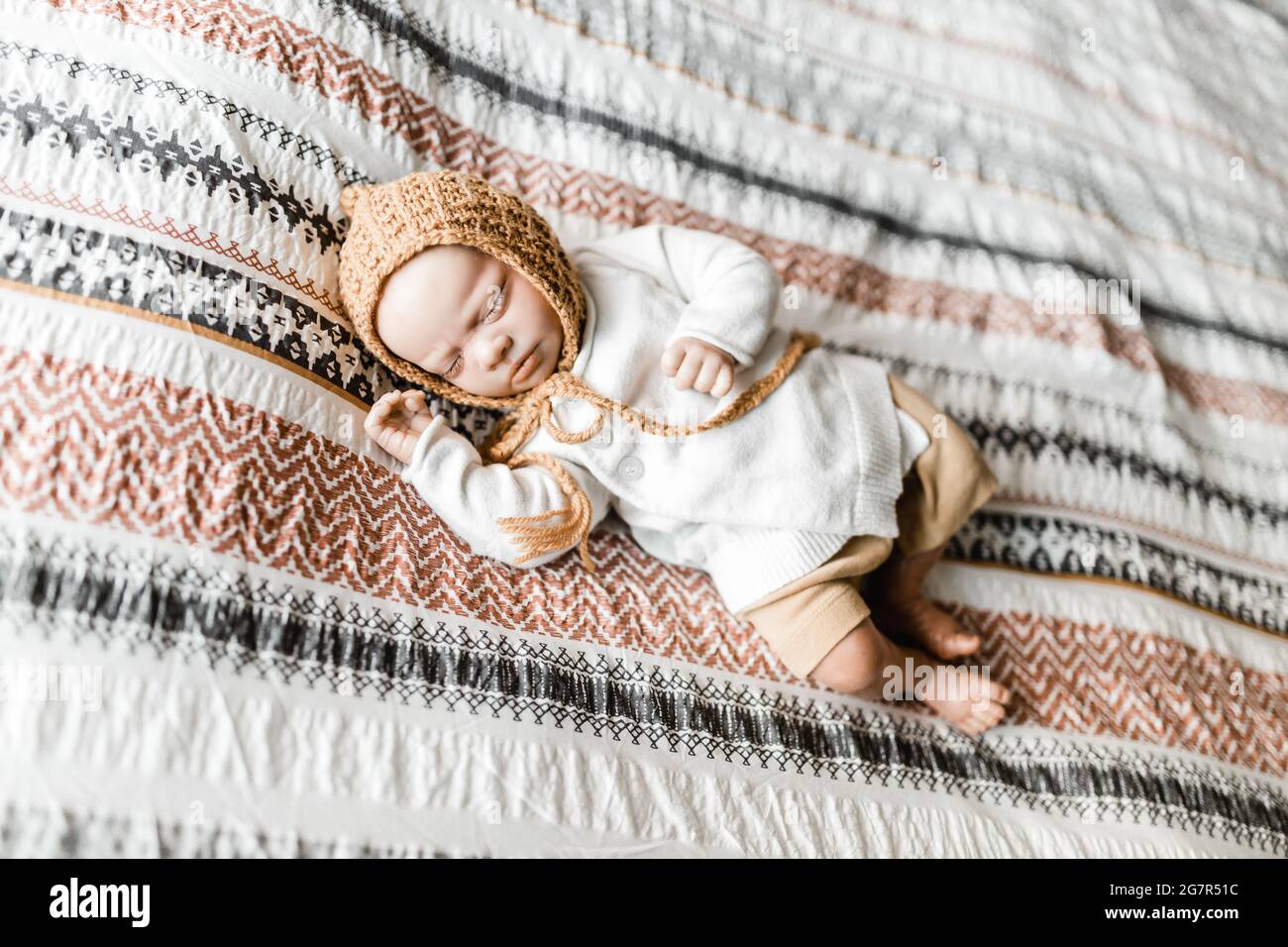 Wiedergeborene Babypuppe, die auf einem Bett mit gemustertem Dovetbezug und einer Haube liegt, Model Release ist nicht erforderlich, da Baby eine lebensechte Puppe ist Stockfoto
