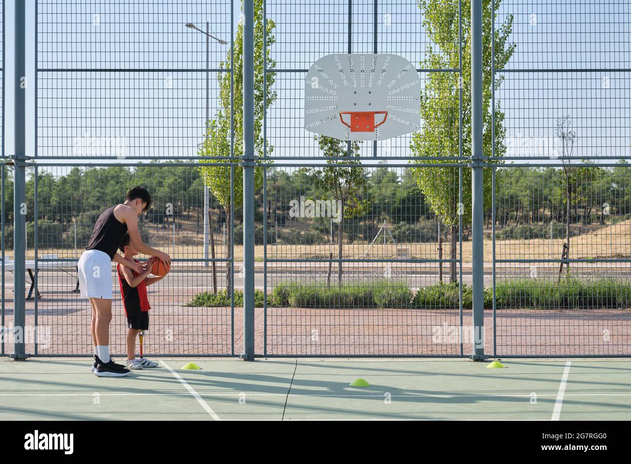 Basketballtrainer zeigt einem Kind mit einer Beinprothese, wie man Basketball schießt. Coach, der ein Kind trainiert. Stockfoto