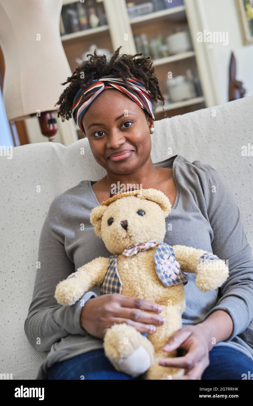 Porträt einer jungen schwarzen Frau mit Afro-Frisur und einem Teddybären im Innenbereich, die auf einem Sofa sitzt. Lifestyle-Konzept. Stockfoto