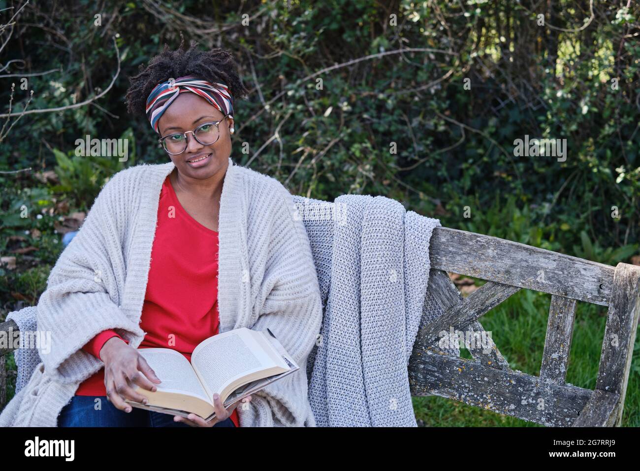 Porträt einer jungen schwarzen Frau mit Afro-Frisur und Brille beim Lesen eines Buches, das auf einer alten Gartenbank sitzt. Lifestyle-Konzept. Stockfoto