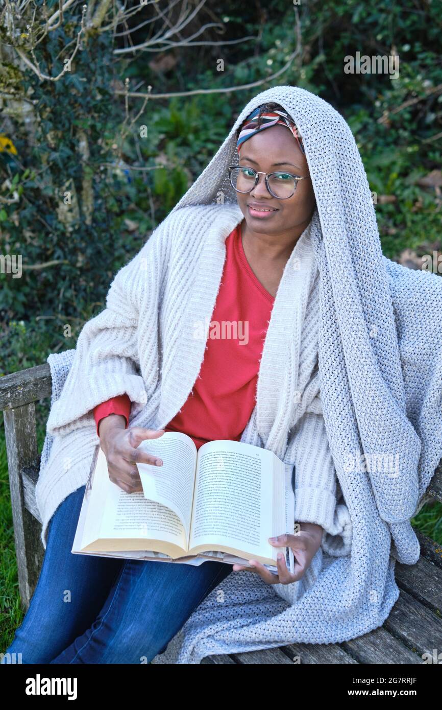 Porträt einer jungen schwarzen Frau mit Afro-Frisur und Brille beim Lesen eines Buches, das auf einer alten Gartenbank sitzt. Lifestyle-Konzept. Stockfoto