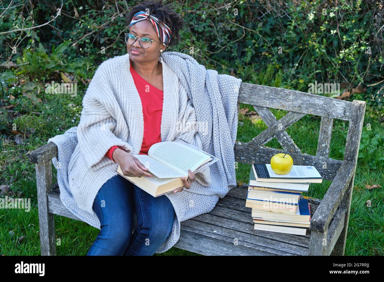 Porträt einer jungen schwarzen Frau mit Afro-Frisur und Brille, die ein Buch liest, das auf einer alten Gartenbank mit einem Bücherstapel sitzt. Lifestyle-Konzept. Stockfoto