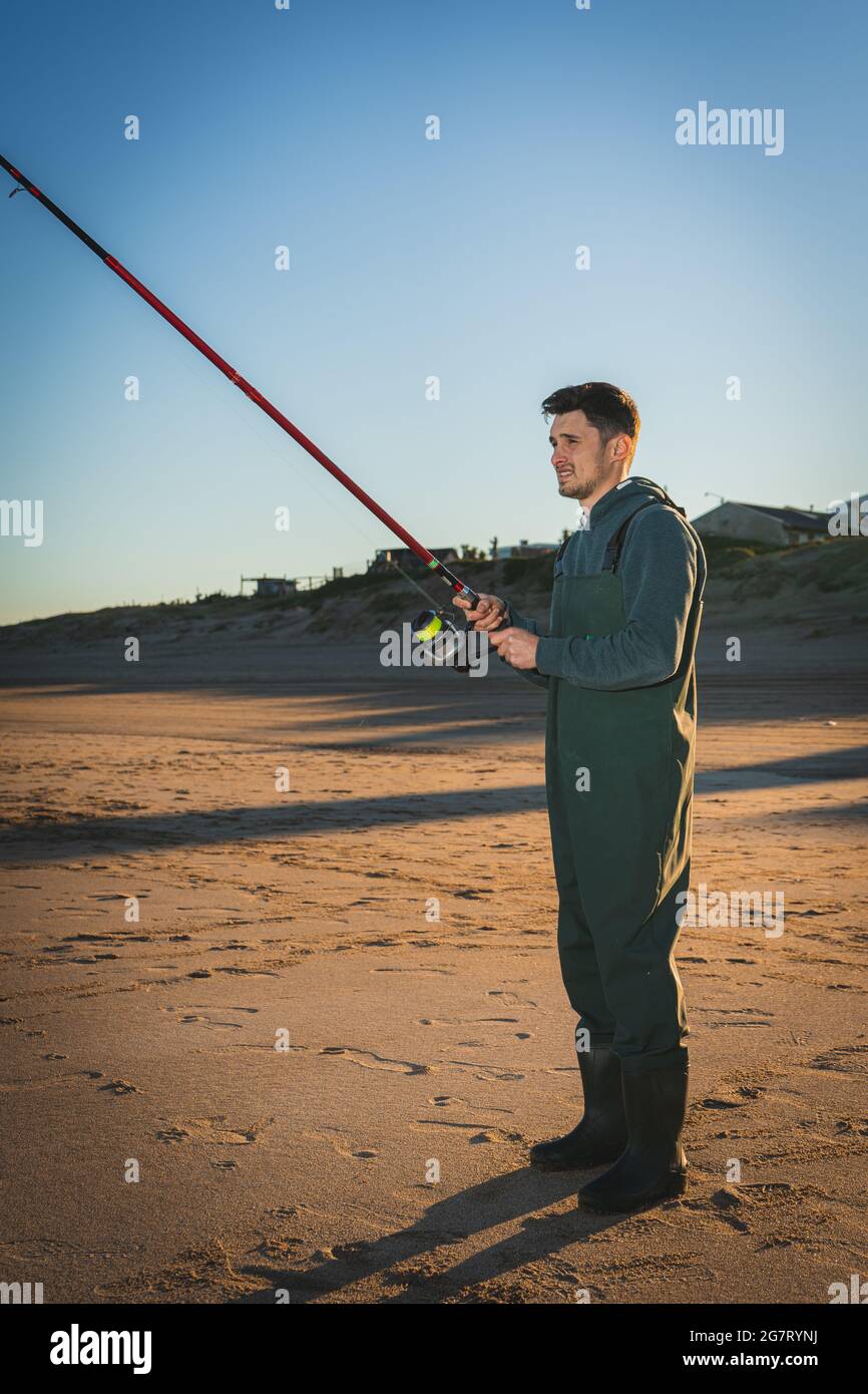 Vertikale Aufnahme eines argentinischen Mannes, der bei Sonnenuntergang am Sandstrand unter einem klaren blauen Himmel fischt Stockfoto