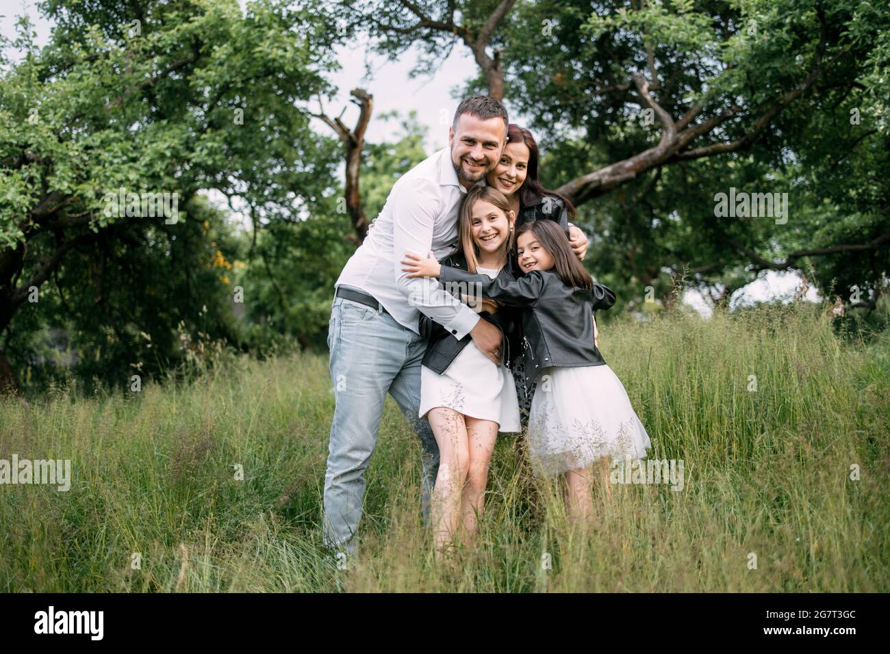 Kaukasische vierköpfige Familie, die in starken Umarmungen im grünen Sommergarten steht. Vater, Mutter und zwei Töchter verbringen die Wochenenden an der frischen Luft. Stockfoto