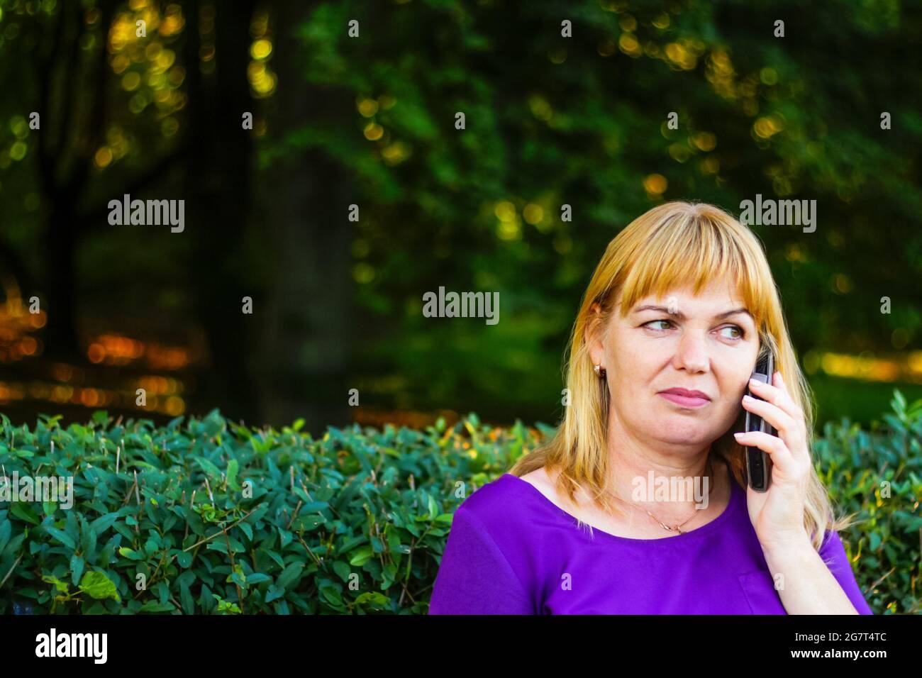 Unschärfe kaukasisch blonde Frau reden, sprechen am Telefon draußen, im Freien. 40-jährige Frau in lila Bluse im Park auf der Bank. Erwachsene Person usi Stockfoto