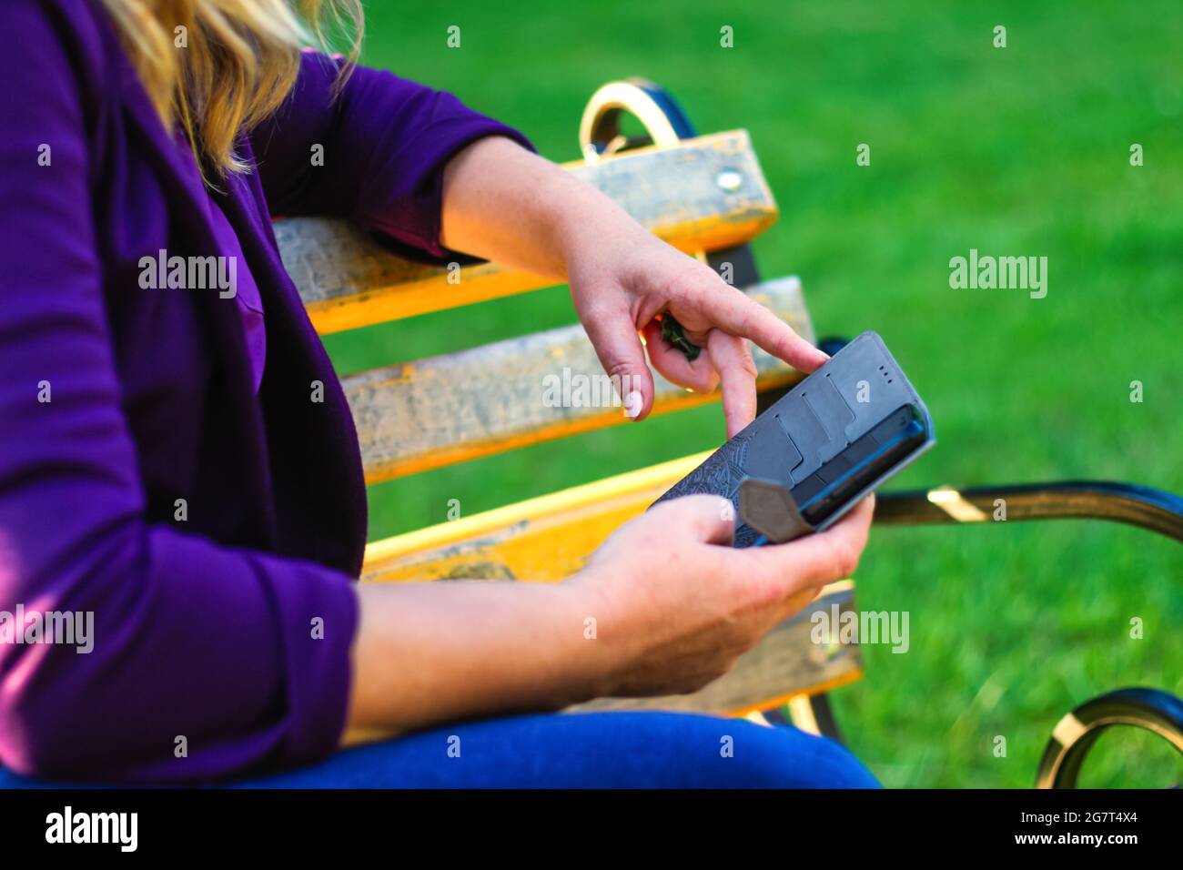 Unschärfe-Unschärfe-Nahaufnahme weibliche Hände suchen auf dem Telefon außerhalb, im Freien. Frau in lila Bluse mit Smartphone, während sie auf einer Bank im Park sitzt. C Stockfoto