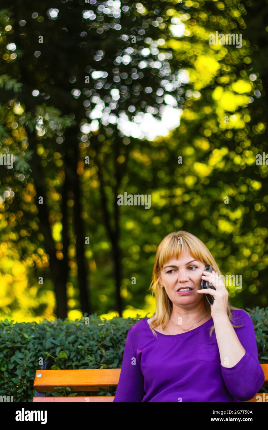 Unschärfe kaukasisch blonde Frau reden, sprechen am Telefon draußen, im Freien. 40-jährige Frau in lila Bluse im Park. Erwachsene Personen, die ein Mobiltelefon verwenden Stockfoto
