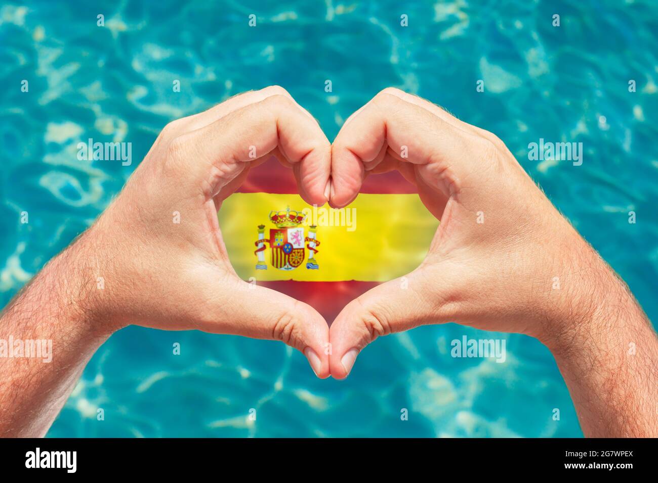 Sommerurlaub in Spanien Konzept. Männliche Hände machen Herzform mit Spanien Flagge im Inneren. Wunderschönes kristallklares türkisfarbenes Wasser mit Ätzstoffen. Stockfoto