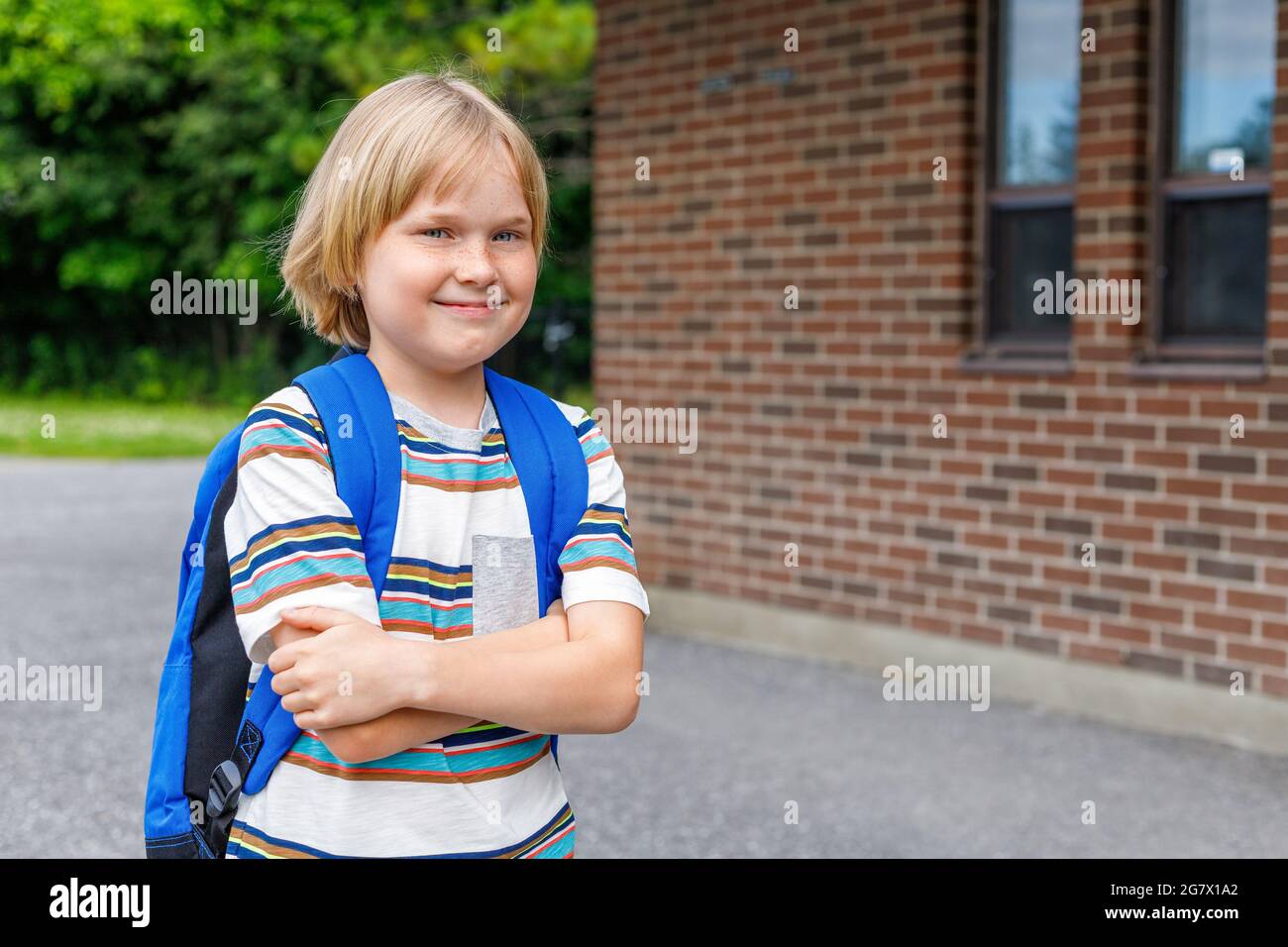 Schönes glückliches Kind in der Nähe des Schulgebäudes auf dem Schulhof. Kleine blonde Studentin mit blauem Rucksack. Zurück zum Schulkonzept. Stockfoto