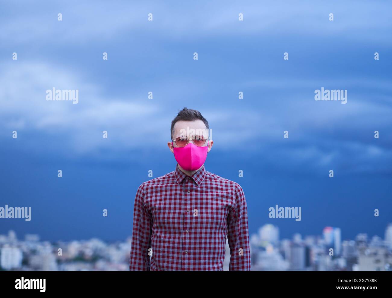 Schöne kaukasische Homosexuell in rosa schützende medizinische Gesichtsmaske suchen gerade in der Kamera. Männliche Person Porträt mit regnerischem Wetter Hintergrund und städtische Skyline. LGBTQ-Themenkonzept. Hochwertiges iimage Stockfoto