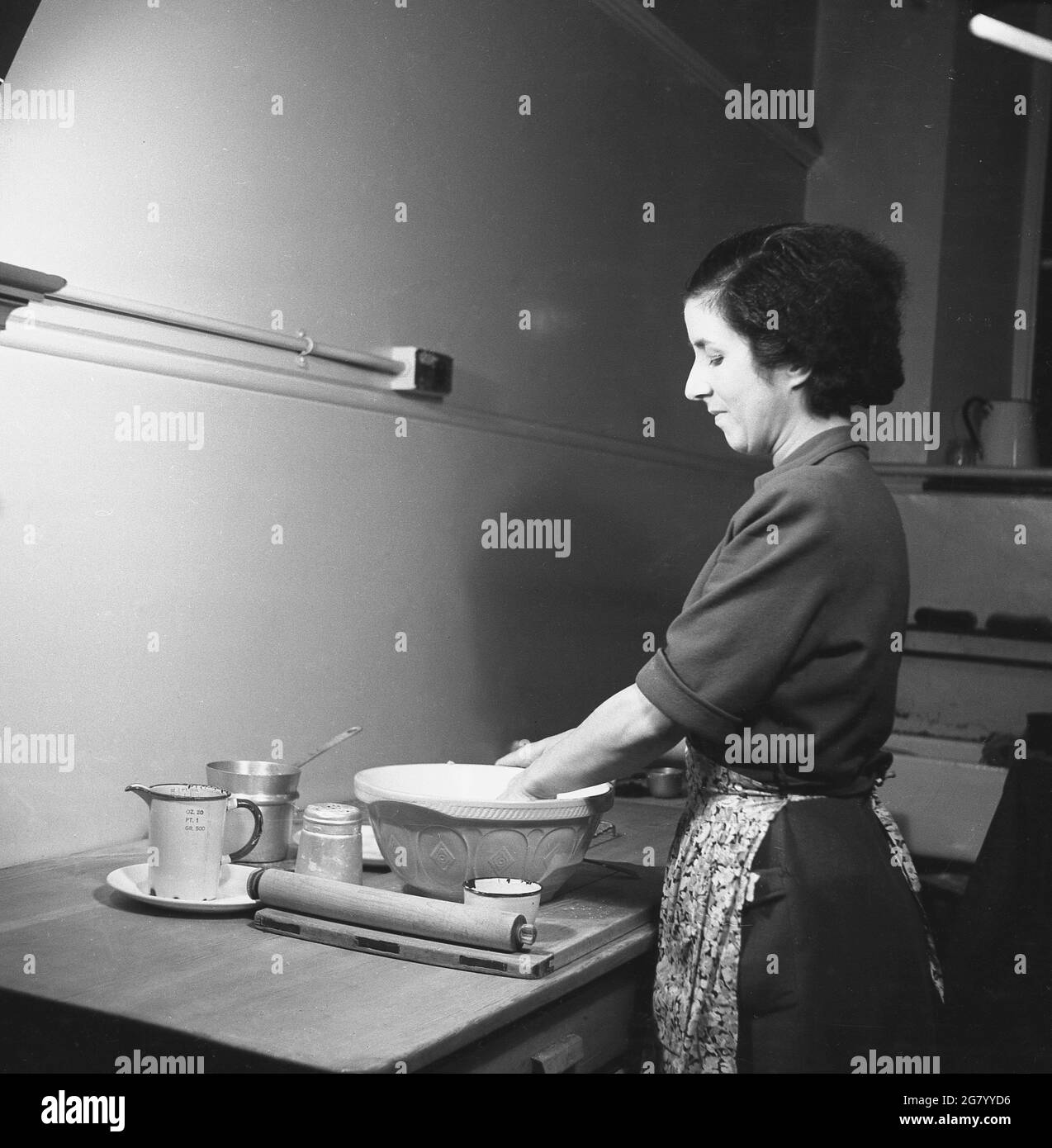 In den 1950er Jahren, historisch, eine Dame in einem Kleid mit einer Schürze, die an einem Holztisch in einer Ecke eines Raumes steht, der Teig in einer Mehlschüssel auf einem hölzernen Gebäckbrett mischt. Mit ihrem Nudelholz neben ihr und wenn sie Teig bereit ist, ist sie bereit, etwas zu backen, England, Großbritannien. Stockfoto