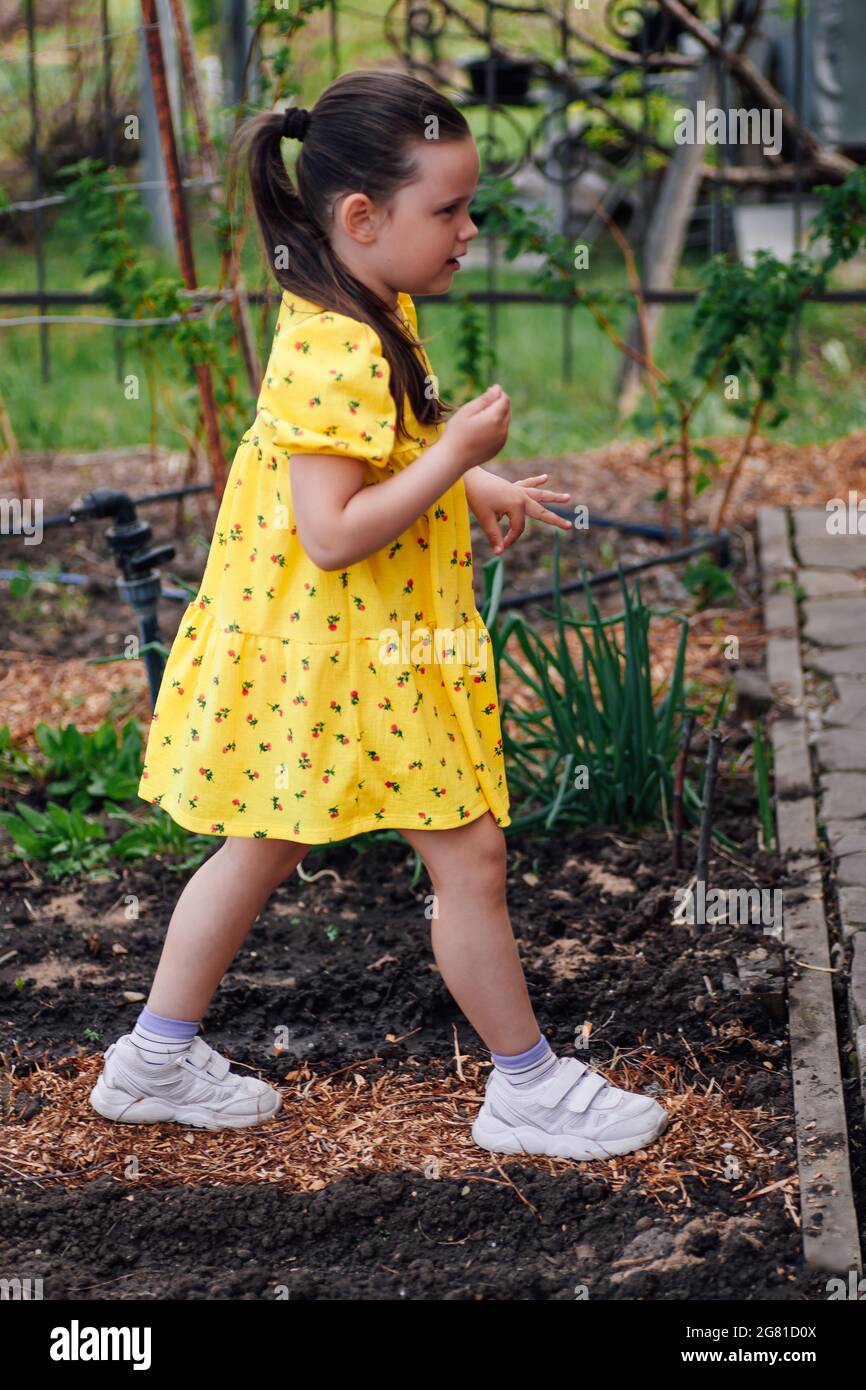 Lifestyle Ganzkörperportrait eines Mädchens in einem gelben Kleid, das zwischen Gemüsebeeten und Pflanzen im Hinterhof läuft, Familienaktivität des Wochenendes Stockfoto