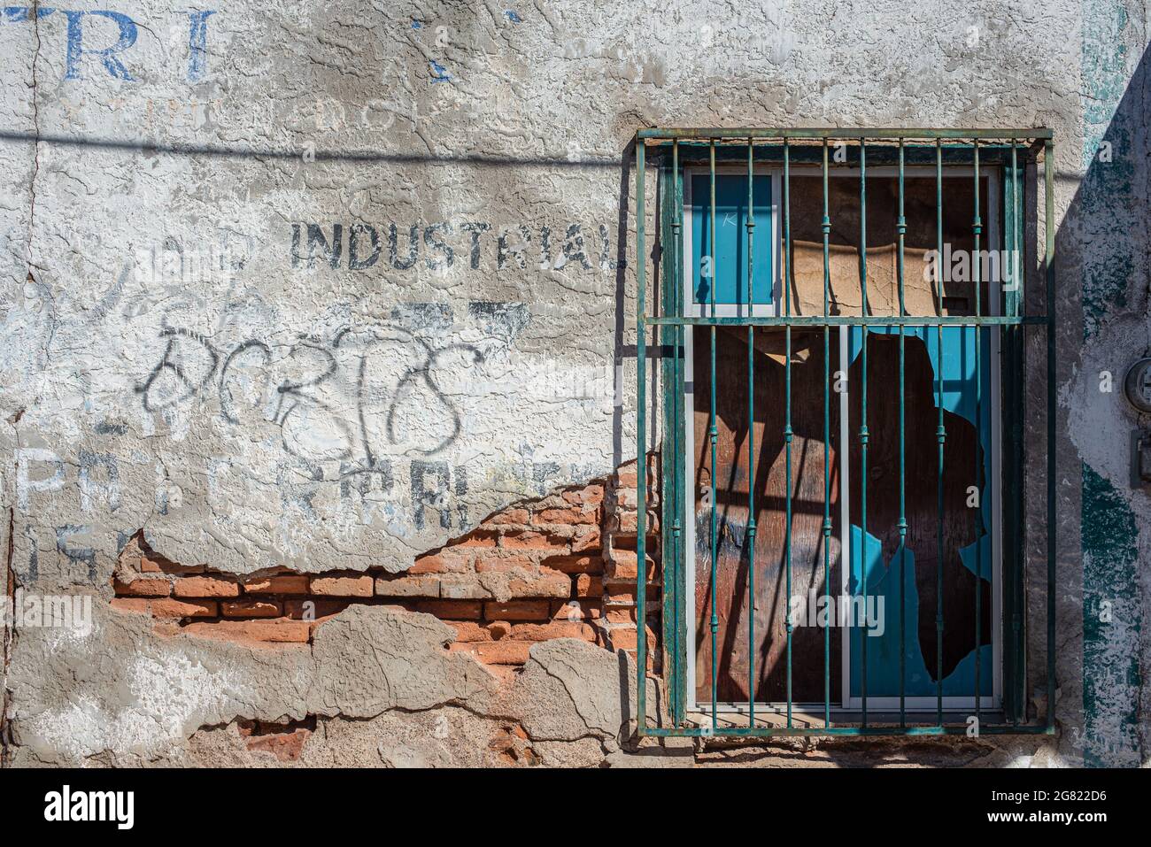 Ein Fenster mit grünen Balken, das Glas gebrochen und leuchtend blauen Flecken in einer bröckelnden weißen Stuckwand mit Ziegelsteinen ausgesetzt. Stockfoto