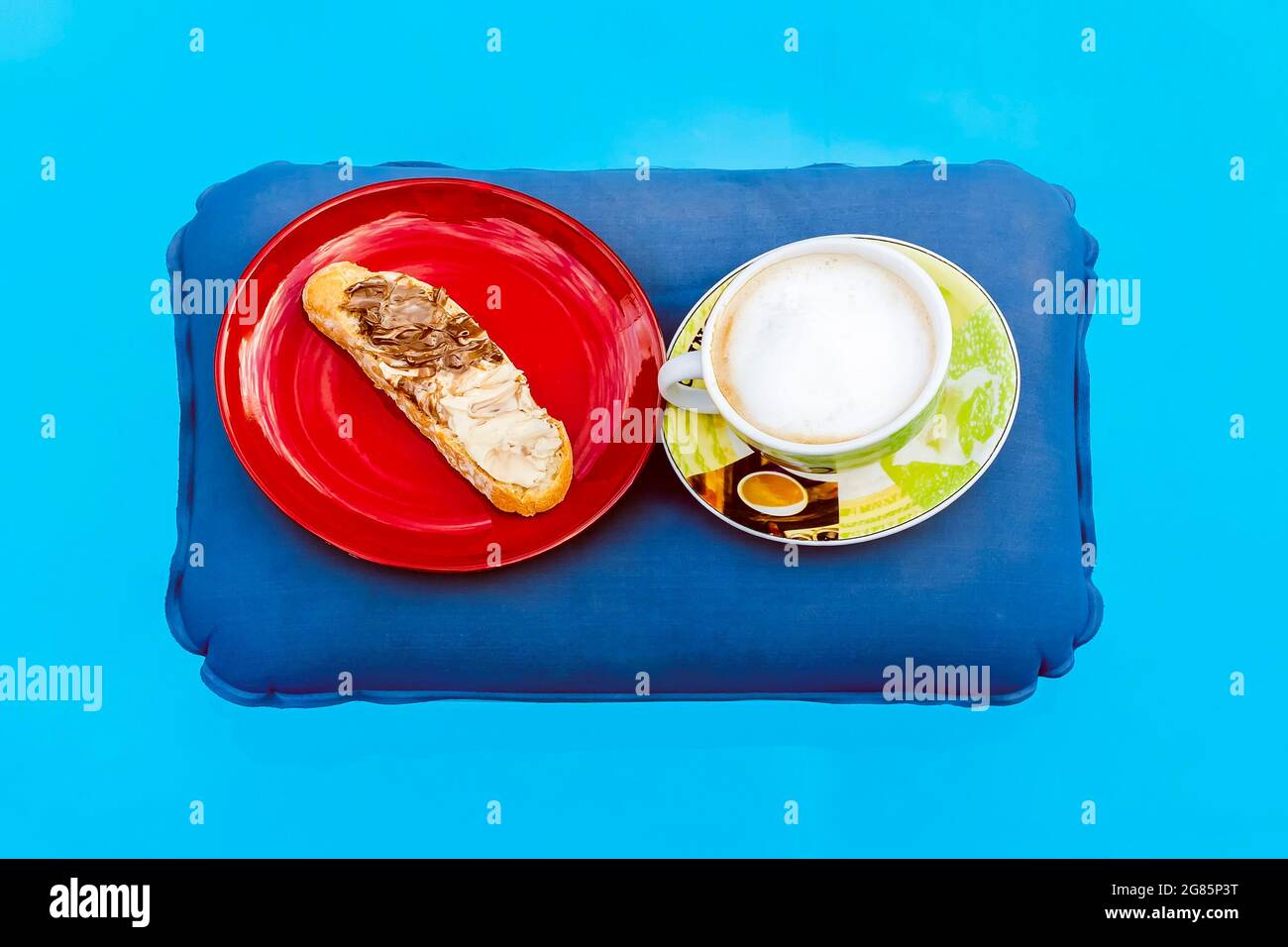 Eine Tasse Cappuccino und ein roter Teller mit einer Scheibe Brot, auf der schwarze und weiße Schokoladencreme auf einem aufblasbaren Kissen aufliegt, das darauf schwimmt Stockfoto
