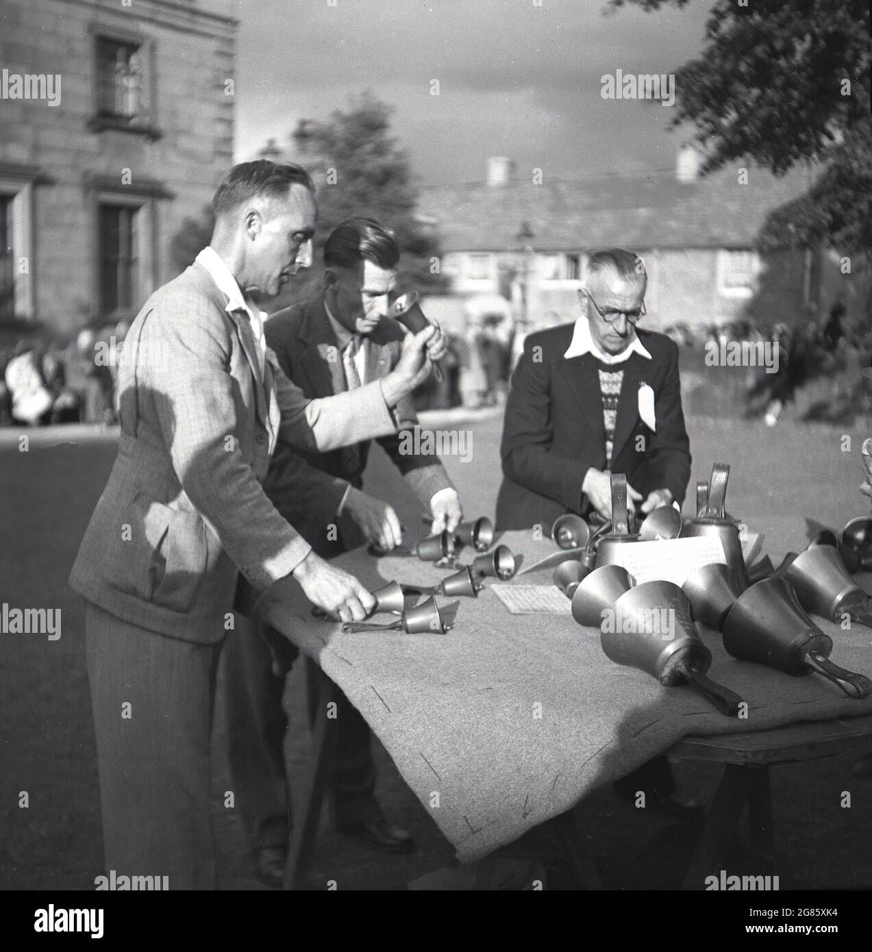 1950er Jahre, historische, Erwachsene Männer, die an einem Tisch stehen und Handglocken prüfen oder testen, England, Großbritannien. Die Brüder Robert & William Cor sollen die Handglocke oder Handglocke zwischen 1696-1724 in Aldbourne, Wiltshire, England, erfunden haben, damit Glockenläuferinnen außerhalb des Turms üben und Proben konnten. Ein Handbell-Team oder -Ensemble ist eine Gruppe von Menschen, die bei Feten und Konzerten musikalische Arrangements läuten. Stockfoto