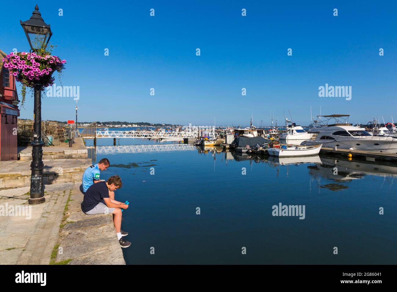 Zwei Jungen, die im Juli am Poole Quay auf einer Mauer sitzen und mit Booten in Poole Harbour, Poole, Dorset, Großbritannien, krabben Stockfoto
