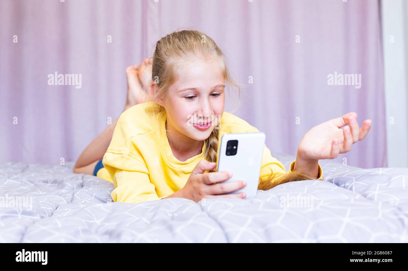 Ein kaukasisches Mädchen im Teenageralter mit einem Smartphone in den Händen, das auf dem Bett liegt, bloggt, mit Freunden in sozialen Netzwerken kommuniziert und kostenlos Geld ausgibt Stockfoto