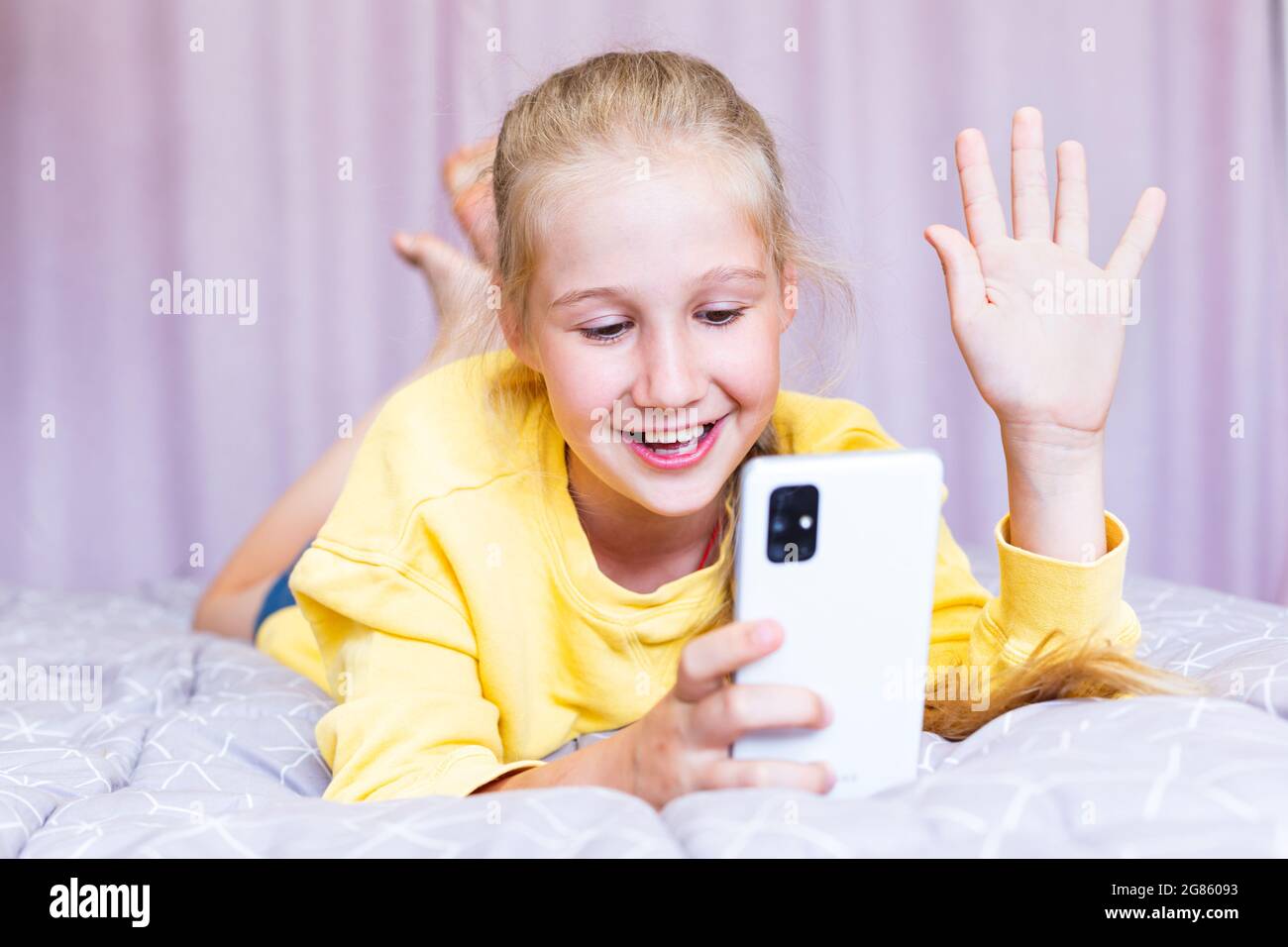 Ein blondes kaukasisches Teenager-Mädchen mit einem Telefon in den Händen, das auf dem Bett liegt, bloggt und in den sozialen Medien mit Freunden in einem gemütlichen Schlafzimmer kommuniziert Stockfoto