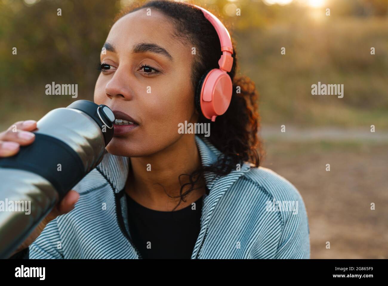 Fokussierte afroamerikanische Sportlerin mit Kopfhörern, die Wasser trinkt, während sie im Park arbeitet Stockfoto
