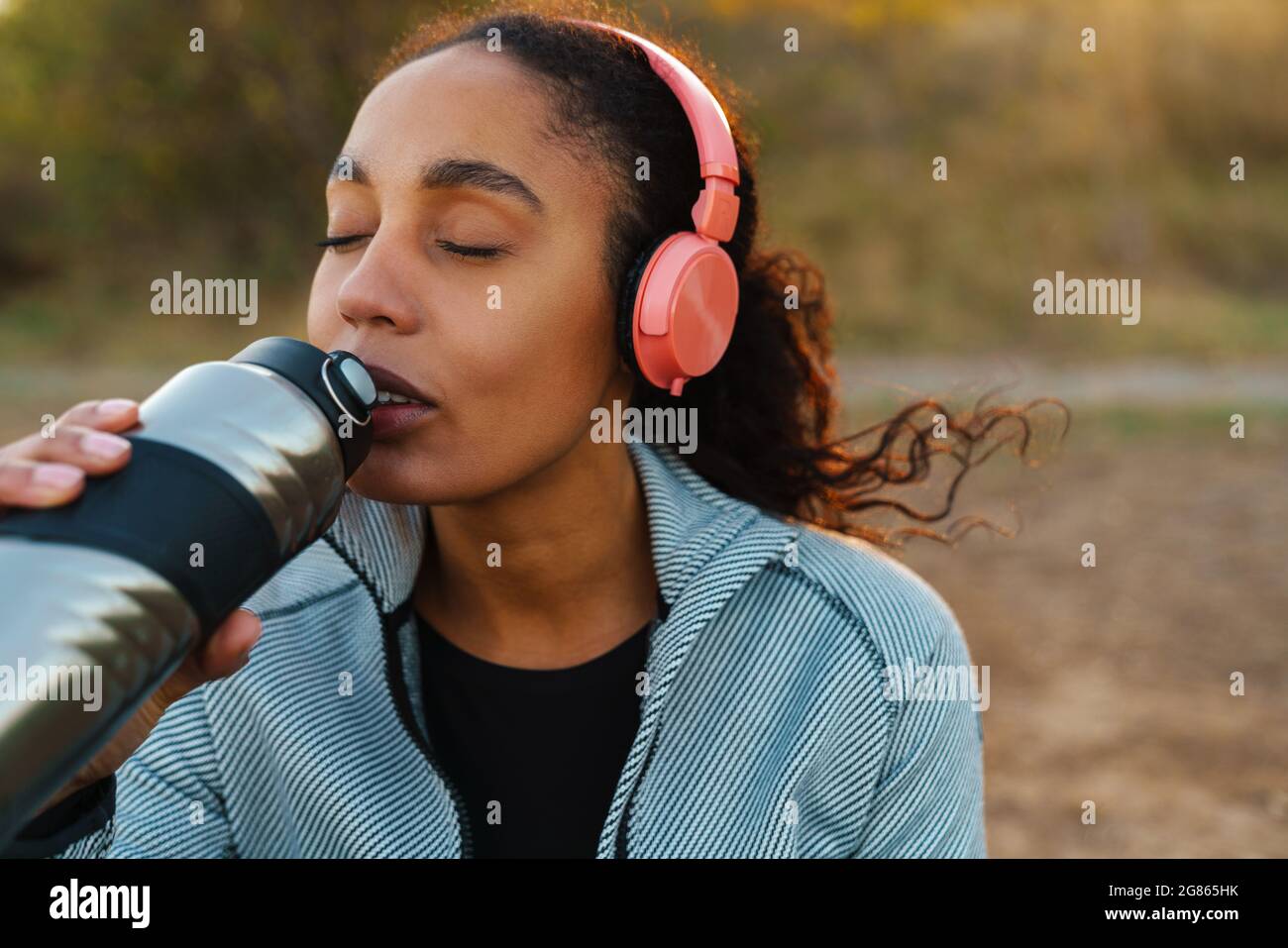 Fokussierte afroamerikanische Sportlerin mit Kopfhörern, die Wasser trinkt, während sie im Park arbeitet Stockfoto