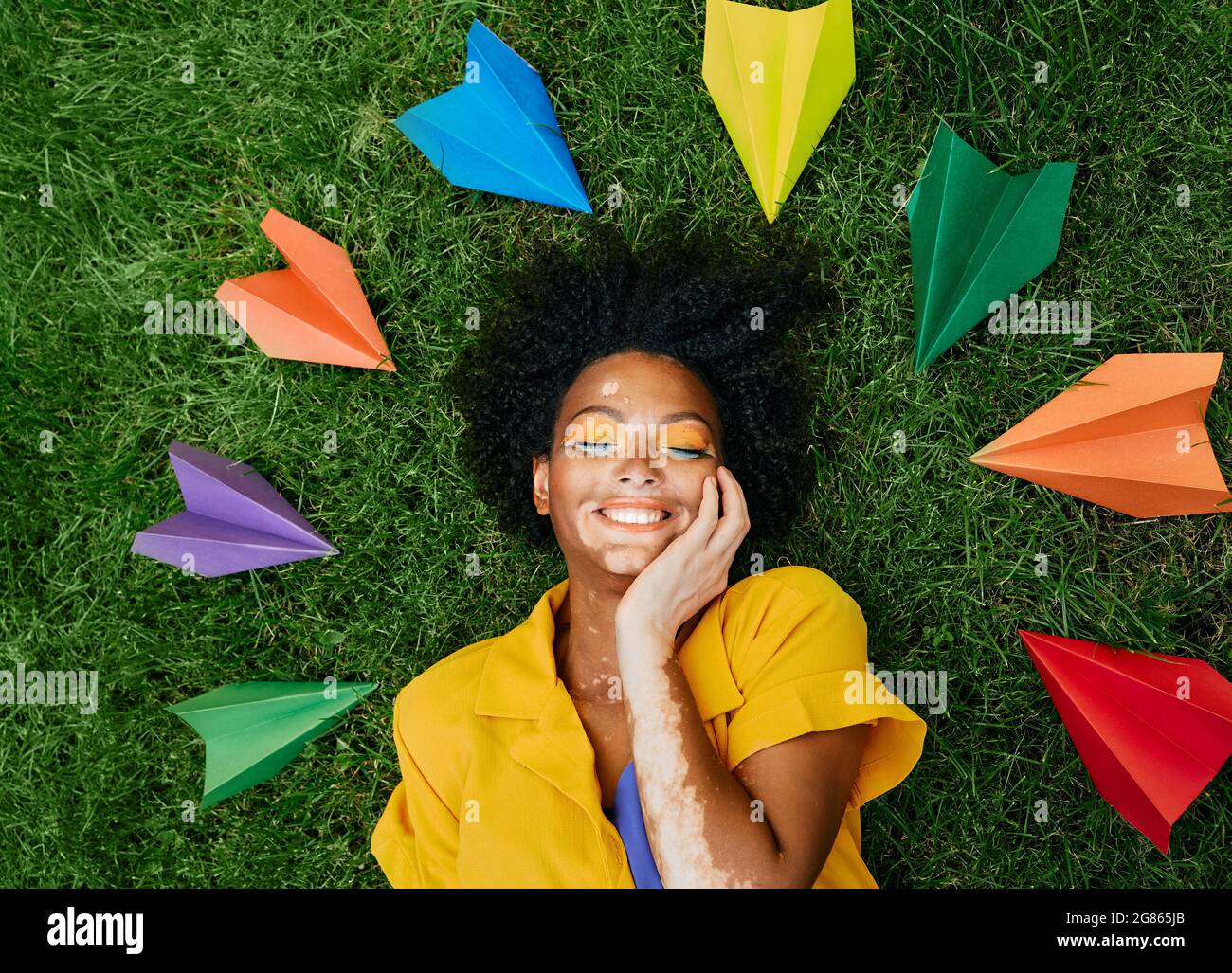 Glückliche Frau, die auf dem grünen Gras liegt und von Urlaub und Reisen träumt, farbige Papierflugzeuge um ihren Kopf. Reisekonzept Stockfoto