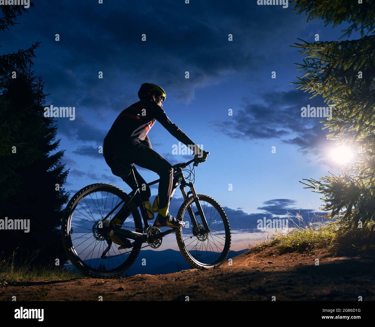 Rückansicht eines Radfahrers unter blauem Abendhimmel mit Wolken. Silhouette des männlichen Radfahrers Fahrrad auf dem Weg in der Nacht Bergwald. Konzept von Sport, Radfahren und aktiver Freizeit. Stockfoto