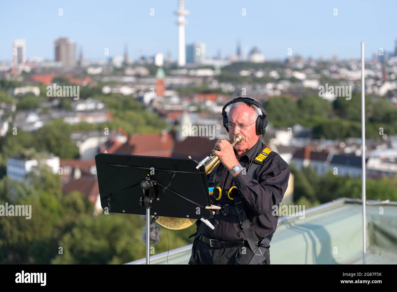 Hamburg, Deutschland. Juli 2021. Ein Hornist spielt sein Instrument auf dem Dach eines Hochhauses vor dem Stadtpanorama mit Fernsehturm. In Zusammenarbeit mit Kampnagel und Elbphilharmonie spielte das Dresdner Sinfonieorchester auf den Dächern der Lenzsiedlung und dem SV Grün-Weiß Eimsbüttel. 500 registrierte Zuschauer konnten im Stadion sitzen. Quelle: Jonas Walzberg/dpa/Alamy Live News Stockfoto