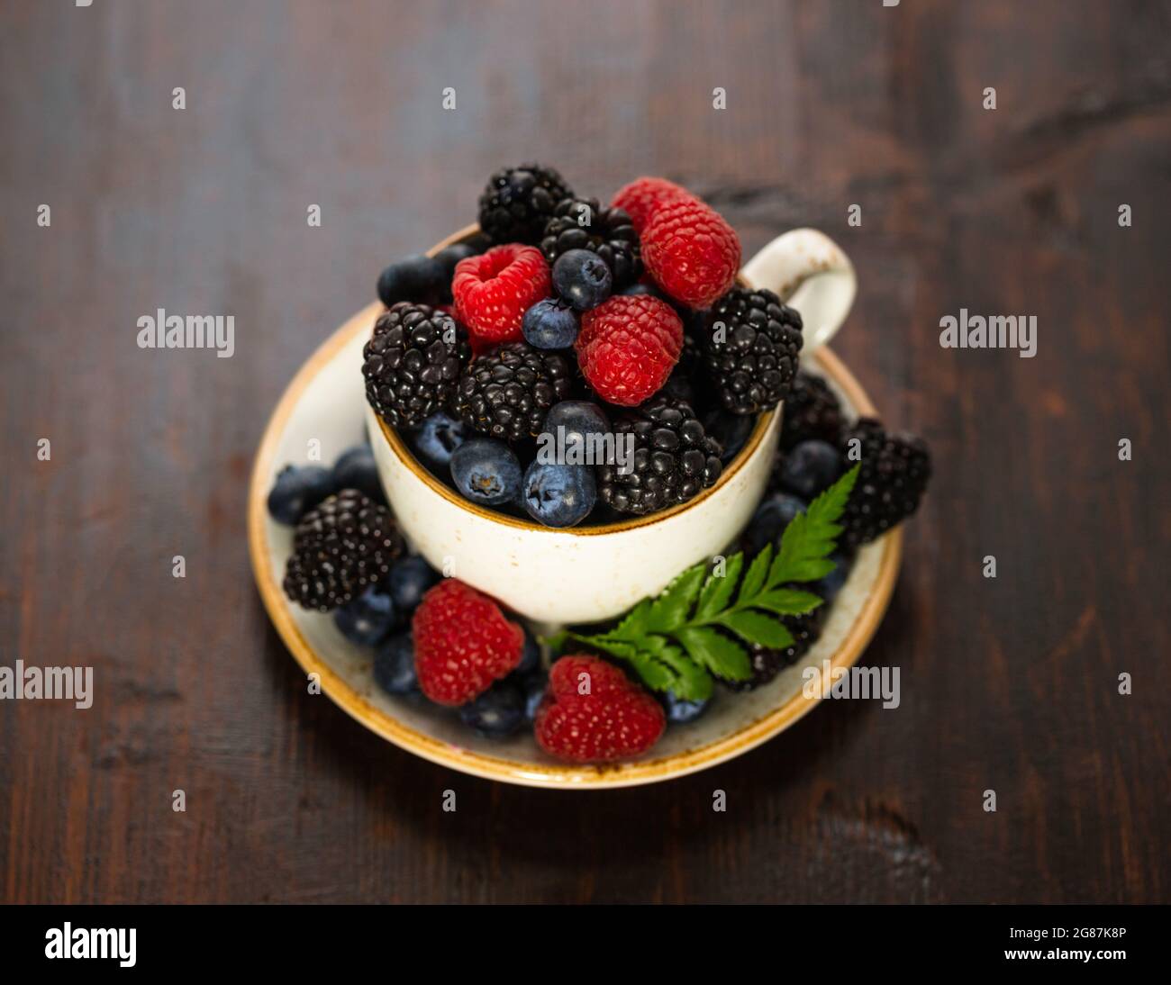 Tasse mit Beeren auf einem hölzernen Hintergrund. Zusammensetzung der Sommerbeere. Stillleben mit Beeren in einer Teetaille. Himbeeren, Brombeeren und Heidelbeeren. Stockfoto