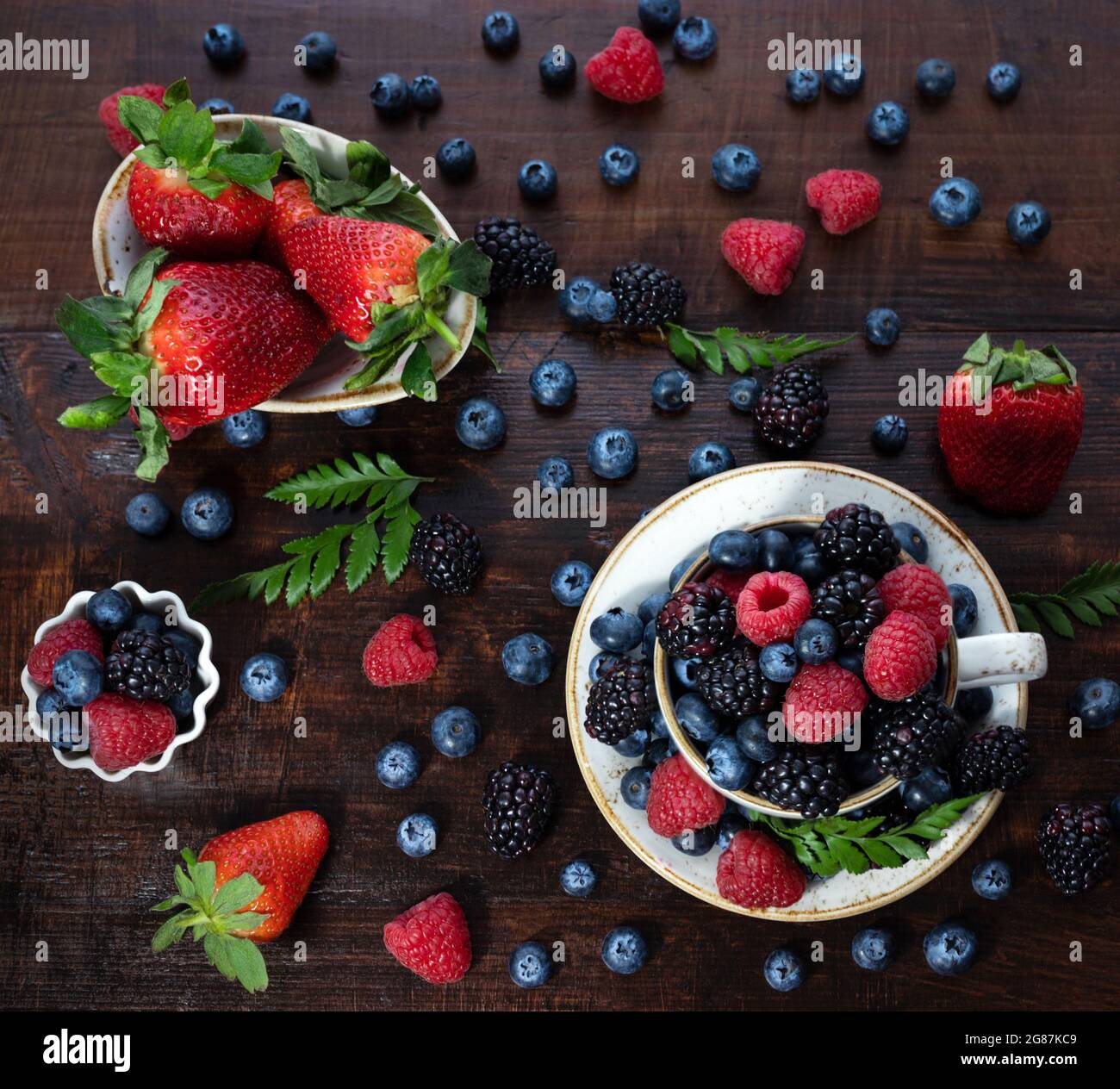 Beerenmischung auf einem hölzernen Hintergrund. Beeren in einer Tasse und Schüssel. Erdbeeren, Blaubeeren, Brombeeren und Himbeeren. Sommerhintergrund. Draufsicht, FLA Stockfoto