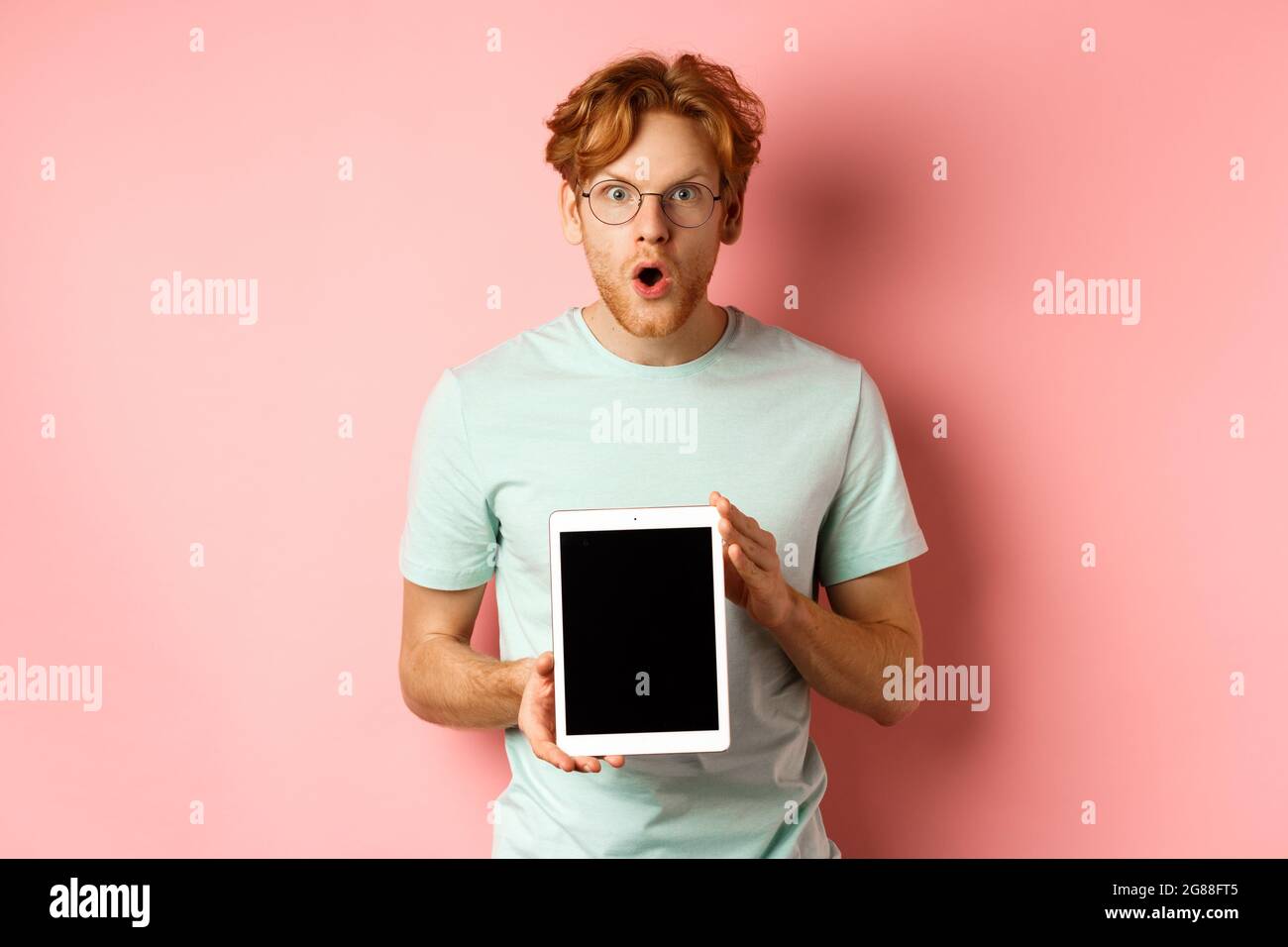 Aufgeregt junger Mann mit roten Haaren und Bart, Check-out Online-Promotion, zeigt digitale Tablet-Bildschirm und starrte auf Kamera erstaunt, über pi stehen Stockfoto