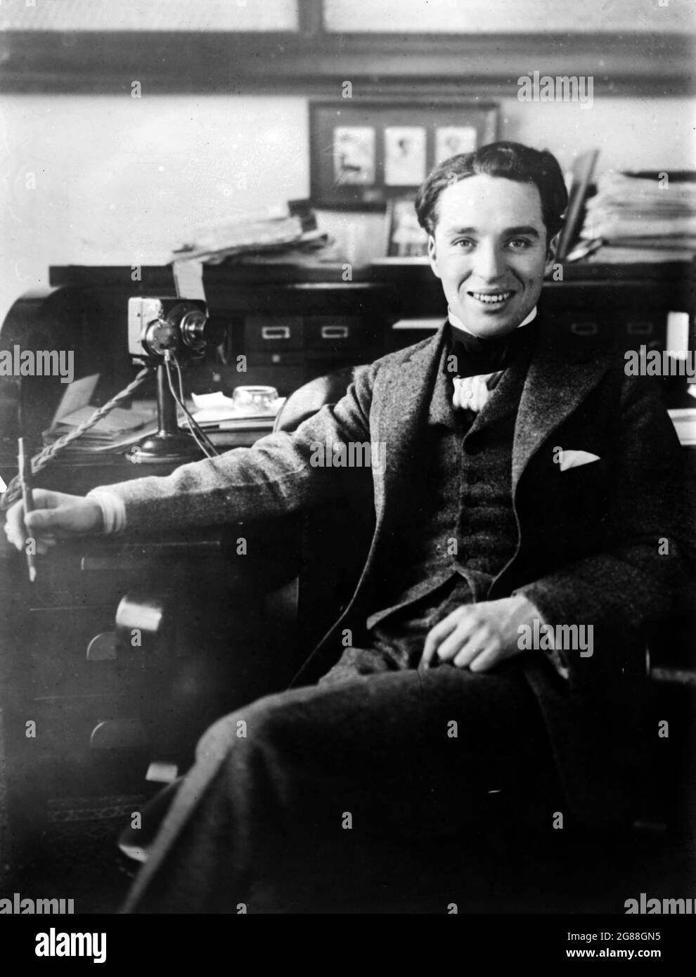 Porträt des Stummfilmstars Charlie Chaplin, um 1915–1920. Chaplin sitzt lächelnd an einem Schreibtisch und sieht entspannt aus. Stockfoto