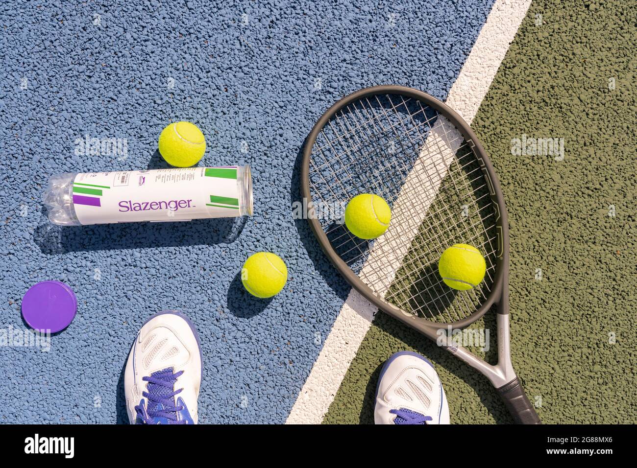 Auf einem harten Tennisplatz mit Tennisschläger und fluoreszierenden gelben Tennisbällen (optisch gelb) blickt man auf die Füße eines Mannes, der Sportschuhe trägt Stockfoto