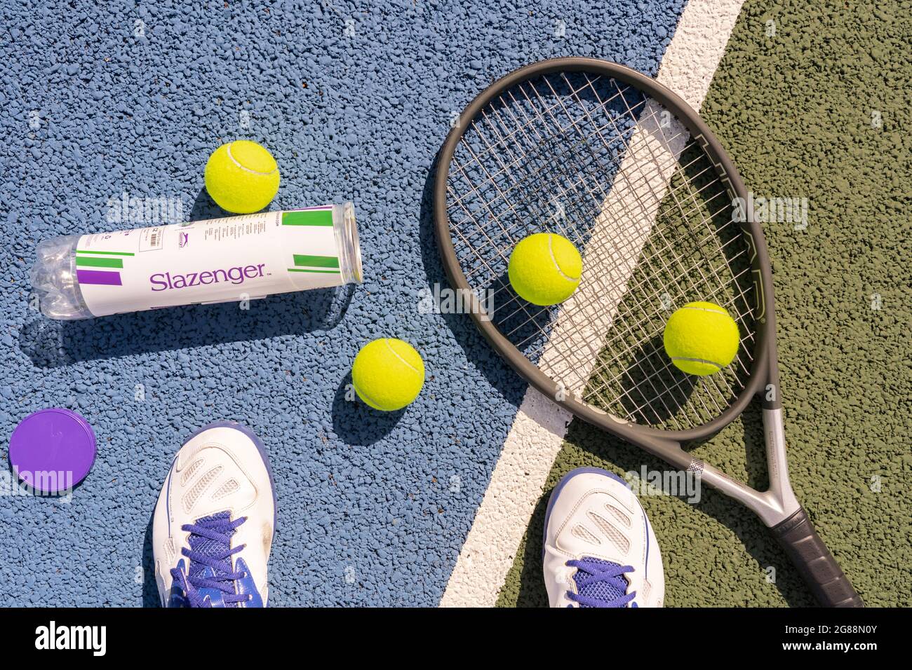 Auf einem harten Tennisplatz mit Tennisschläger und fluoreszierenden gelben Tennisbällen (optisch gelb) blickt man auf die Füße eines Mannes, der Sportschuhe trägt Stockfoto