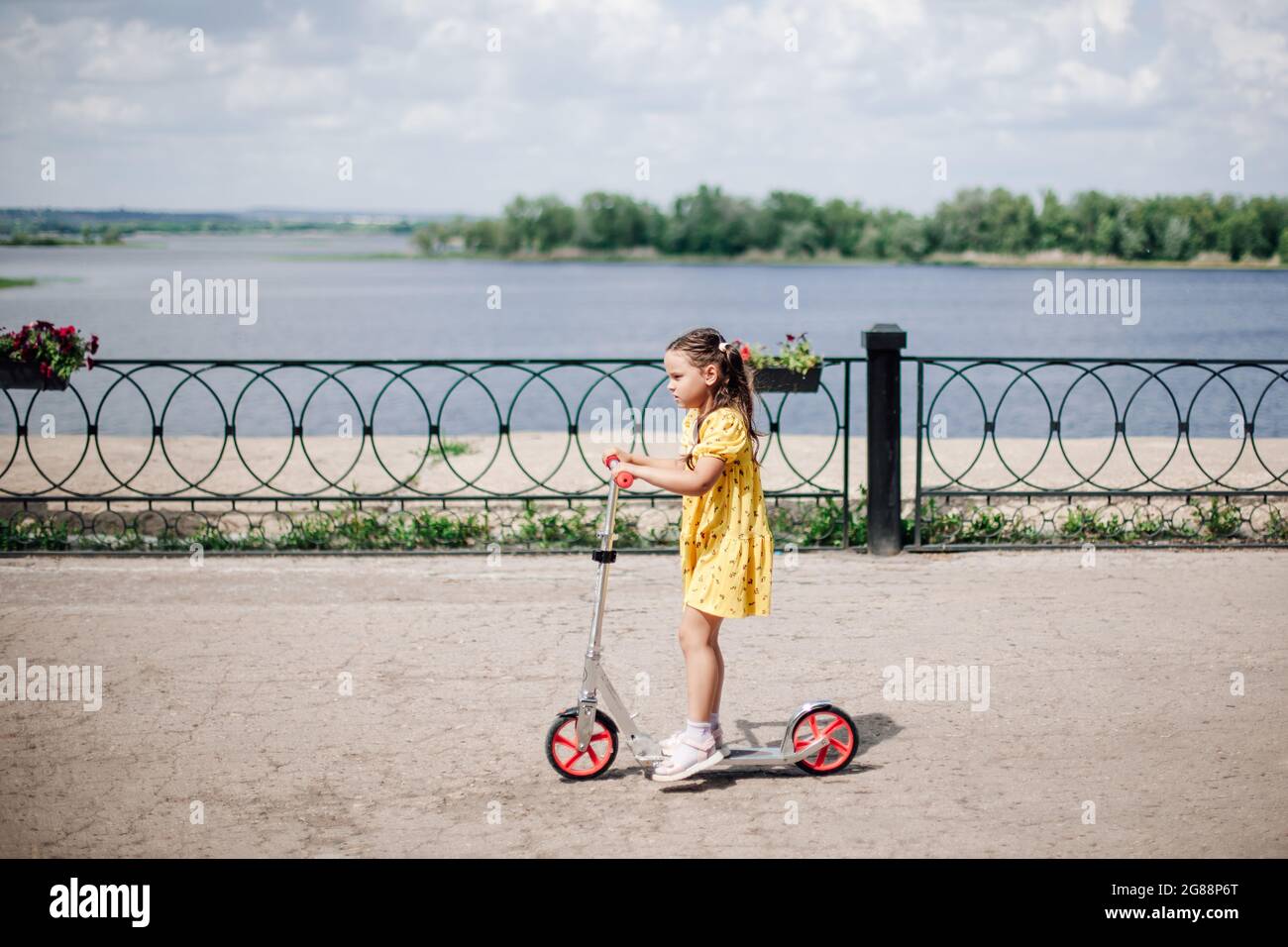 Lifestyle-Porträt, der Moment der Bewegung eines Mädchens auf einem Roller fährt ein Mädchen in einem Kleid mit einem Motorroller mit einem gusseisernen Zaun entlang des Flussufers Stockfoto